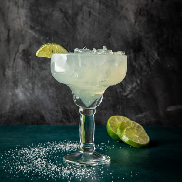 A margarita in a glass with ice and a lime wedge, surrounded by salt and lime slices.