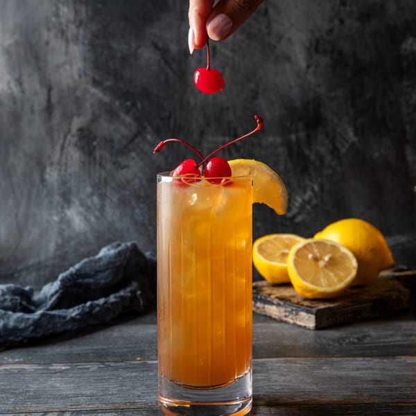 A hand garnishes a tall glass of iced tea with a cherry, alongside lemons on a wooden board.