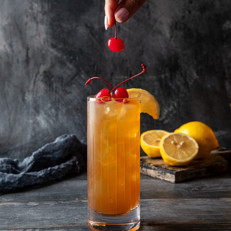 A hand garnishes a tall glass of iced tea with a cherry, alongside lemons on a wooden board.