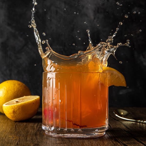 A Tom, Dick & Hank cocktail splashes from a glass with lemons and a spoon on a wooden table.